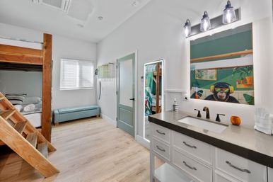 Bright, modern guest room with wooden bunk bed and ladder, teal bench under a window, mint-green door, and a white vanity with dark countertop; mirror reflects a playful colorful monkey mural wearing headphones.