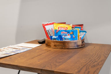 Round wooden tray of assorted packaged snacks (Lay's chips, mini Oreos and cookies) on a wooden table with papers and a remote against a gray wall.