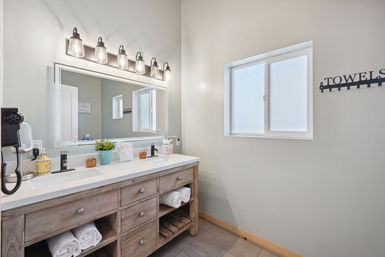 Bright modern bathroom with natural wood double-sink vanity and white quartz countertop, large mirror topped by five industrial-style glass lights, rolled white towels in open shelves, frosted window and wall-mounted towel rack