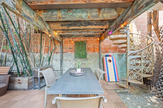 Cozy urban backyard patio with a long concrete dining table and wicker chairs under weathered wooden beams, spiral wrought-iron staircase, bamboo plants, striped cornhole board and string lights against an exposed brick wall.