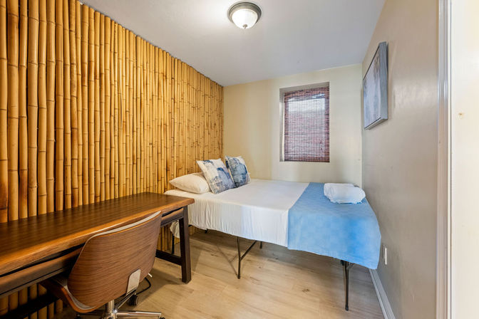 Cozy compact bedroom with bamboo accent wall, twin bed dressed in white linens and blue throw, patterned pillows, wooden desk and chair, bamboo window shade and light wood flooring.