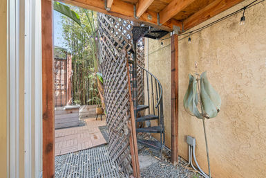 Cozy urban backyard nook under a wooden deck featuring a black metal spiral staircase, diagonal lattice screen, bamboo planter, paver patio with gravel floor, string lights and textured stucco wall.