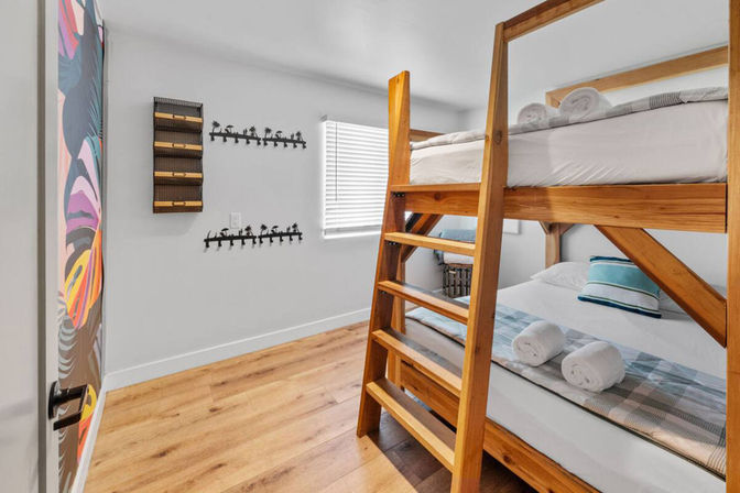 Bright coastal-style bedroom with wooden bunk beds and ladder, rolled towels on the lower bed, palm-tree wall hooks, colorful door mural, and light hardwood floor