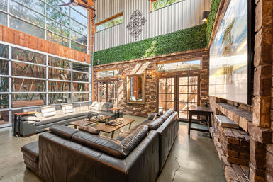 Sun-drenched industrial loft living room with floor-to-ceiling glass garage door, large leather sectional, exposed brick walls, indoor green wall and modern coffee tables.