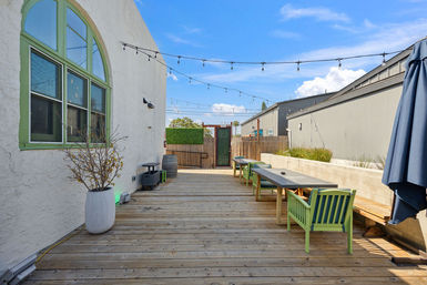 Sunny urban wooden deck patio with hanging string lights, communal picnic tables and lime-green chairs beside a white stucco building with a green-trim arched window.