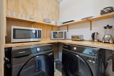 Compact laundry-kitchenette: black front-load washer and dryer under a wooden countertop with a microwave, toaster oven, two-slice toaster, electric kettle and blender, and stacked plates and glasses on open wood shelves.