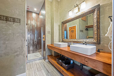 Spa-like modern-rustic bathroom with wooden double-sink vanity and vessel sinks, ornate framed mirror, textured plaster walls, tile floor and glass walk-in shower with dark tiles.