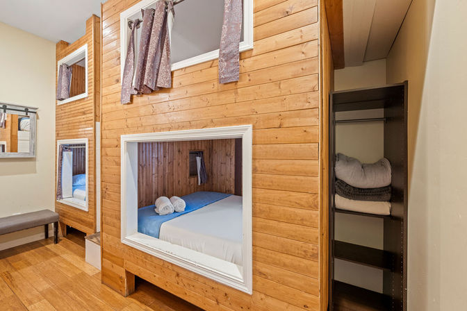 Cozy wooden pod bunk room with recessed sleeping cubbies, blue-and-white bedding and rolled towels, curtained windows, mirror and bench.
