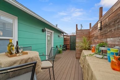 Cozy urban backyard deck with turquoise house siding, wooden privacy fence, bistro table with wine glasses, colorful planters and potted plants under a blue sky