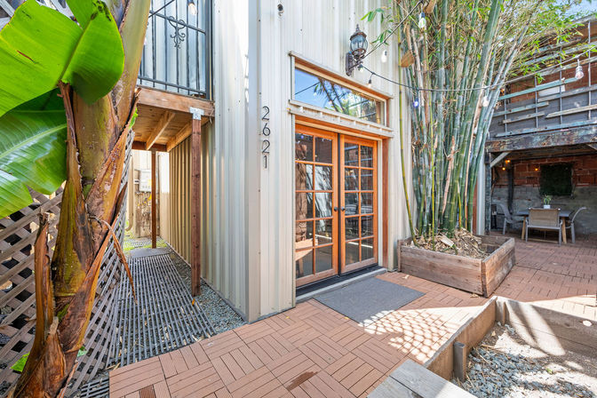 Sunlit urban courtyard patio with wooden French doors in a metal-clad building, raised bamboo and banana planters, string lights overhead, interlocking wood-look tiles and a cozy outdoor seating nook.