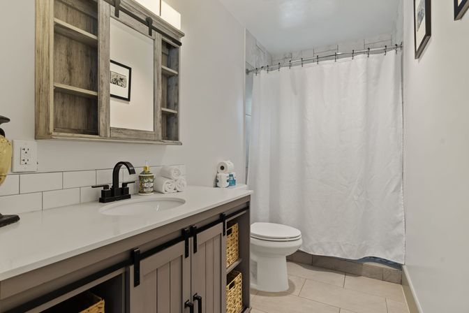 Cozy modern farmhouse bathroom with gray wood vanity and wicker baskets, white quartz countertop, matte black faucet, rustic mirrored cabinet, subway tile backsplash, white shower curtain over tub, toilet, and neutral tile floor.