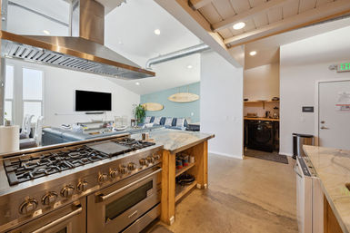 Bright open-concept coastal kitchen and living room with stainless-steel professional range and hood, marble-topped island, concrete floors, blue sectional beneath decorative surfboards on a teal accent wall, wall-mounted TV, large windows, exposed ductwork and a tucked laundry nook.