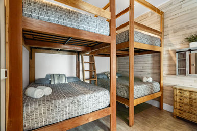 Cozy rustic bunk room with two stacked wooden double beds (four sleeping spaces), gray striped bedding, rolled white towels, ladder, whitewashed shiplap wall and small dresser.