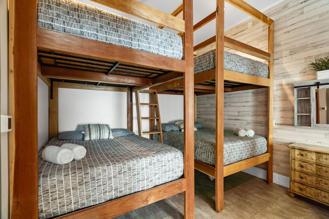 Cozy rustic bunk room with two stacked wooden double beds (four sleeping spaces), gray striped bedding, rolled white towels, ladder, whitewashed shiplap wall and small dresser.