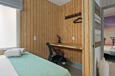 Cozy wood-paneled coastal cottage bedroom with a single bed in a mint-green throw and rolled white towels, a compact wall-mounted desk with a black office chair and coat hooks, doorway revealing a beach-themed mural in the next room.