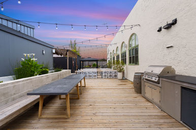 Outdoor wooden deck patio at sunset with long communal table, string lights overhead, buffet grill and seating by arched windows — casual al fresco dining and event space.