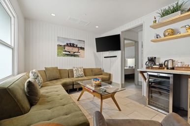 Sunlit modern living room in a rental suite with an olive-green sectional, glass coffee table with snacks and magazines, wall-mounted TV, kitchenette area with coffee maker and wine fridge, white shiplap walls and decorative shelves.
