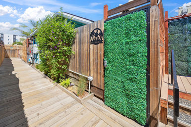 Sunny beachy boardwalk entrance with wooden decking, a wood fence and a green faux-boxwood gate beside flowering shrub under a bright blue sky.
