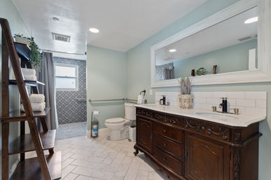 Spa-like bathroom with carved dark wood double vanity and large mirror, pale aqua walls, herringbone floor tile, ladder towel shelf, grab bar, and patterned walk-in shower.