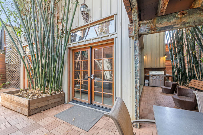 Cozy urban courtyard patio with tall bamboo planters, wooden French doors, wicker seating, tiled deck and stainless steel grill.