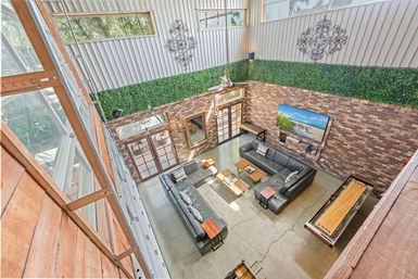 Loft-style industrial living room seen from mezzanine: high ceiling with metal and brick walls, faux green hedge trim, two gray leather sectionals around wooden coffee tables, wall-mounted flat-screen TV, shuffleboard table, concrete floor and large windows/French doors filling the open-concept space with natural light.