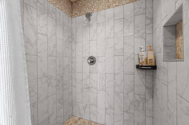 Modern walk-in shower with vertical gray marble-look tiles, pebble stone border and floor, chrome showerhead and control, black shelf with bottles and recessed niche