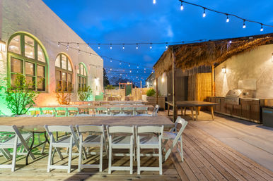 Outdoor wooden-deck patio at dusk with long rustic tables and white folding chairs, twinkling string lights, arched stucco windows and a thatched-roof outdoor kitchen with stainless grill