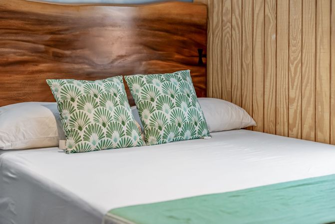 Cozy bedroom with smooth wooden headboard, white linens and two green palm-print throw pillows on a neatly made bed against a vertical wood-paneled wall