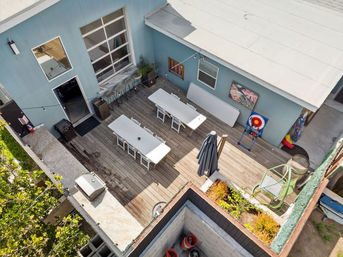 Aerial view of a sunlit rooftop patio with wooden decking, two long white dining tables with benches, bar stools along a blue exterior wall, string lights, patio umbrella, potted plants and a colorful outdoor target game.