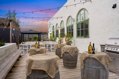 Sunset outdoor patio with string lights, wooden deck, wine barrels draped in burlap used as cocktail tables, white folding chairs under a gazebo, stainless-steel grill and a white stucco building with green arched windows — cozy al fresco dining scene.