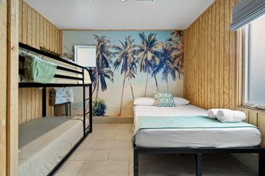 Cozy beach-themed bedroom with palm tree wall mural, wood-paneled walls, black metal bunk bed and double bed with rolled towels and window light