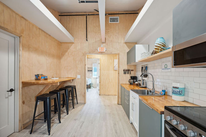 Cheerful loft-style kitchenette with light wood-paneled walls, butcher-block counters and white subway tile backsplash; stainless stove and microwave, sink and coffee station on right, wooden breakfast bar with four black metal stools on left and a doorway with an exit sign ahead.