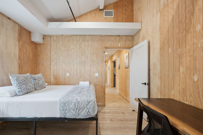 Light wood-paneled bedroom interior with a neatly made bed, patterned throw and pillows, simple wooden desk and open doorway leading to a hallway