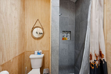 Cozy wood-paneled bathroom with a white toilet, round rope-hung mirror, gray tiled walk-in shower with a built-in niche holding shampoo bottles, and a floral-accent shower curtain.