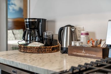 Modern kitchen countertop coffee station with drip coffee maker, stainless steel electric kettle, coffee grinder, wicker tray of filters and pods, disposable cups and creamers on a marble surface, ready for morning brew