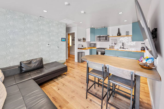 Bright modern open-plan apartment with teal kitchen cabinets, white subway tile backsplash, wooden island with bar stools, black leather sectional, patterned accent wall, and light hardwood floors.