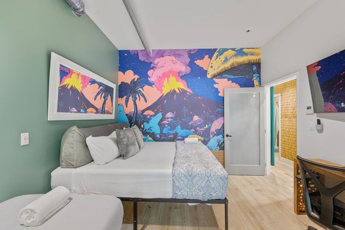 Bright modern boutique bedroom with a large bed, playful neon tropical volcano mural with palm trees and oversized mushrooms, green accent wall, desk and open bathroom door — colorful vacation rental interior.