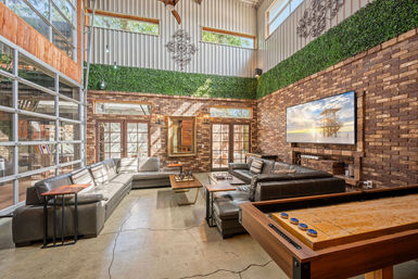 Sunlit industrial loft living room with high ceilings, exposed brick and faux greenery wall, leather sectional, glass garage-door windows, wall-mounted TV and shuffleboard table.