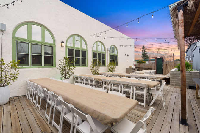 Al fresco wooden-deck patio with long banquet tables covered in burlap, white folding chairs, string lights overhead, and arched green-trim windows on a stucco wall at sunset
