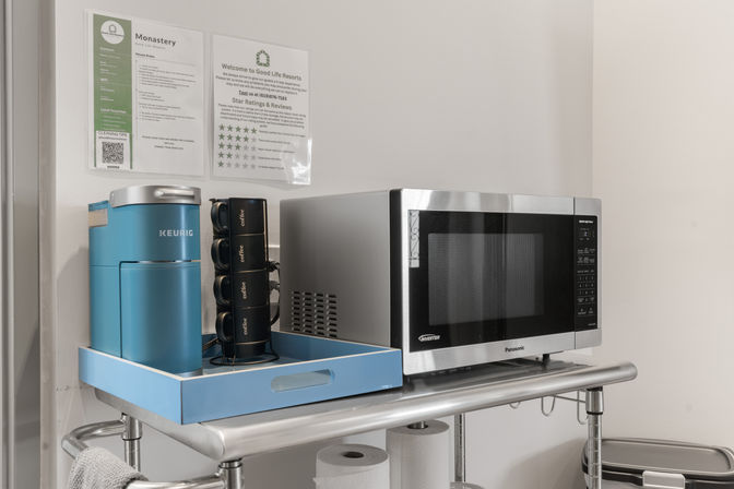 Compact hotel kitchenette shelf with stainless-steel microwave, bright blue single-serve coffee maker and stacked black mugs on a blue tray, with paper towels stored below