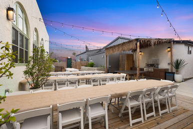 Outdoor patio dining on a wooden deck with long banquet tables draped in burlap, rows of white folding chairs, hanging string lights at sunset and a thatched-roof bar with grill and potted plants.