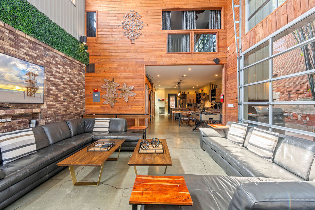 Bright urban industrial loft living room with high wood-paneled wall, exposed brick and green wall, large gray leather sectional sofas, wooden coffee tables, garage-style glass wall, and open-concept kitchen in the background.