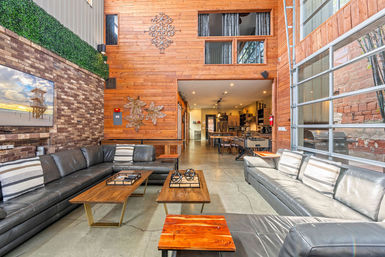Bright urban industrial loft living room with high wood-paneled wall, exposed brick and green wall, large gray leather sectional sofas, wooden coffee tables, garage-style glass wall, and open-concept kitchen in the background.