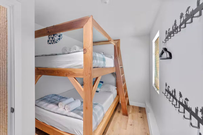 Cozy beach-cottage bunk bedroom with sturdy pine bunk beds, blue plaid bedding, rolled towels, palm-tree wall hooks, white walls, light wood floor and a narrow sunlit window.