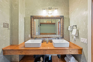 Rustic double bathroom vanity with two white vessel sinks on a wooden countertop, ornate framed mirror, wall sconce lighting, mounted hair dryer and towel against textured plaster walls.