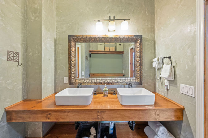 Rustic double bathroom vanity with two white vessel sinks on a wooden countertop, ornate framed mirror, wall sconce lighting, mounted hair dryer and towel against textured plaster walls.