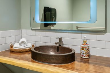 Copper hammered vessel sink on a wooden countertop with chrome faucet, white subway-tile backsplash, decorative soap dispenser and rolled towels in a wicker tray — modern spa-inspired bathroom vanity.
