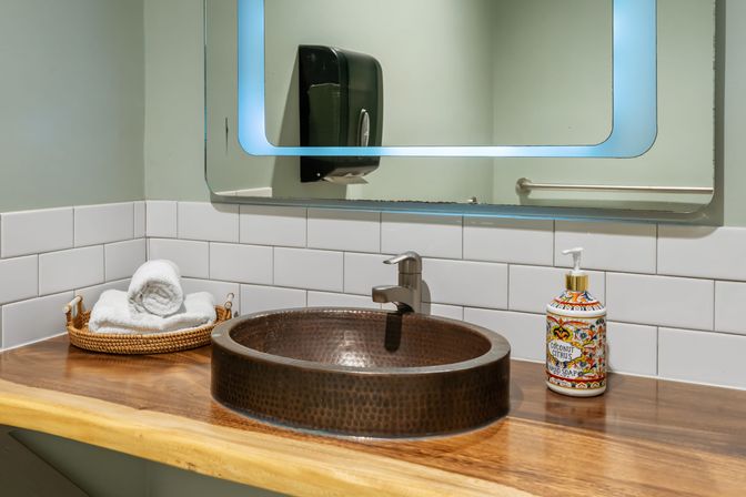 Copper hammered vessel sink on a wooden countertop with chrome faucet, white subway-tile backsplash, decorative soap dispenser and rolled towels in a wicker tray — modern spa-inspired bathroom vanity.