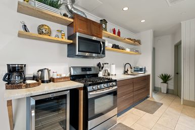 Bright modern kitchen with stainless-steel gas range and microwave, warm wood floating shelves, marble countertop, cozy coffee station, mini wine fridge and potted plant