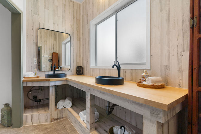 Bright rustic bathroom vanity with long light-wood countertop, two black round vessel sinks and matte black faucets, frosted window, rectangular mirror, open shelving with rolled white towels and wicker tray of toiletries.